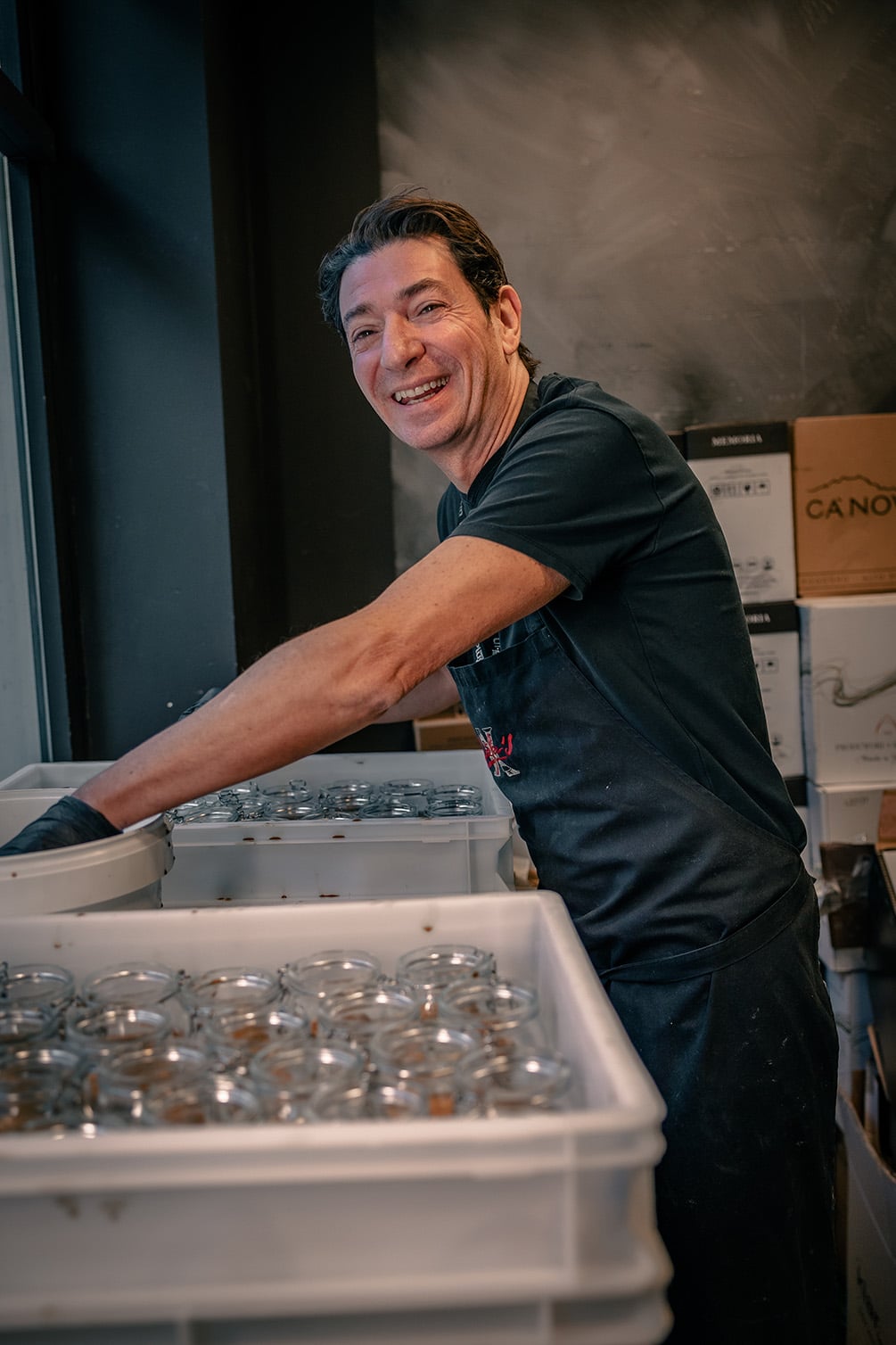 Smiling Stefano in a black apron stands beside plastic bins filled with glass jars, suggesting a busy workspace. The atmosphere is industrious and cheerful.