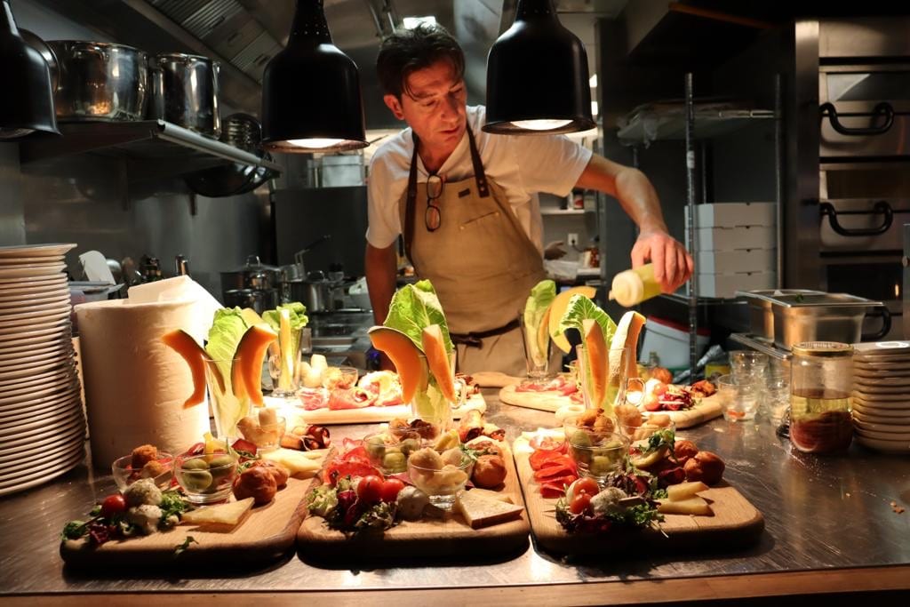Chef in a restaurant kitchen garnishes elaborate dishes with fresh vegetables and colorful ingredients on wooden boards under warm lighting. Preparing snacks for a company meeting.