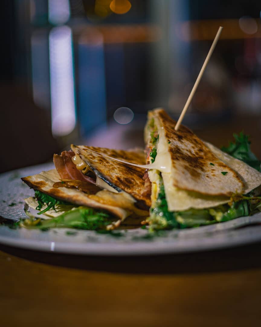 Grilled sandwich with melted cheese, greens, and meats, cut into halves on a plate. A toothpick pierces the sandwich. Warm, inviting ambiance.