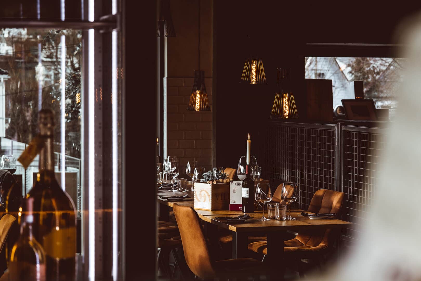 Cozy restaurant interior with dim lighting, featuring a set dining table with brown chairs, wine bottles, and a lit candle, creating a warm, inviting ambiance.